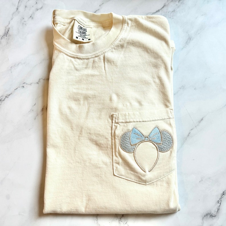 Op de afbeelding: Een cr&egrave;mekleurig T-shirt met een zakje aan de linkerkant. Het zakje heeft een geborduurd ontwerp van een lichtblauwe strik en oren.