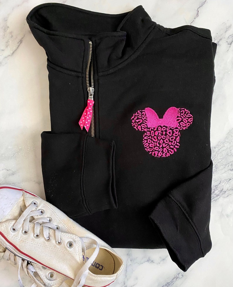 May include: Black sweatshirt with a pink and white leopard print Minnie Mouse design on the front. The sweatshirt has a zipper up the front and a pink and white polka dot zipper pull.