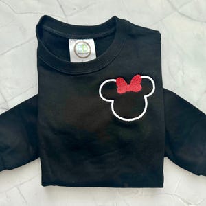 Op de afbeelding: Zwart sweatshirt met lange mouwen met een witte omtrek van een muizenkop en een rode strik. De crewneck-trui heeft geribde manchetten. Een klein label is zichtbaar bij de halslijn. Het kledingstuk is netjes opgevouwen.