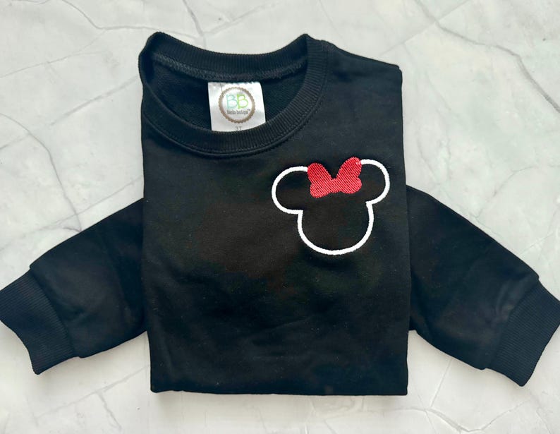 Op de afbeelding: Zwart sweatshirt met lange mouwen met een witte omtrek van een muizenkop en een rode strik. De crewneck-trui heeft geribde manchetten. Een klein label is zichtbaar bij de halslijn. Het kledingstuk is netjes opgevouwen.