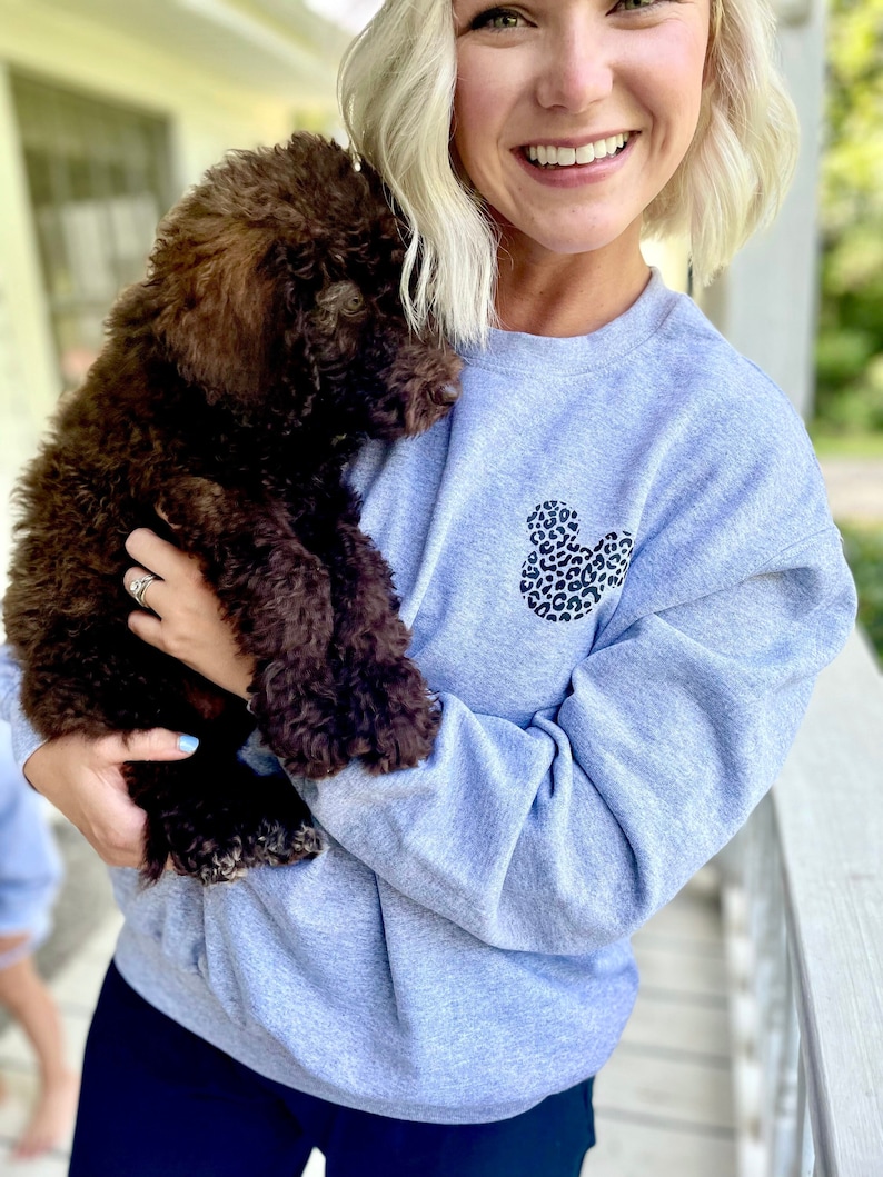 May include: A woman wearing a light gray sweatshirt with a black and white leopard print Mickey Mouse design. She is holding a brown puppy in her arms.