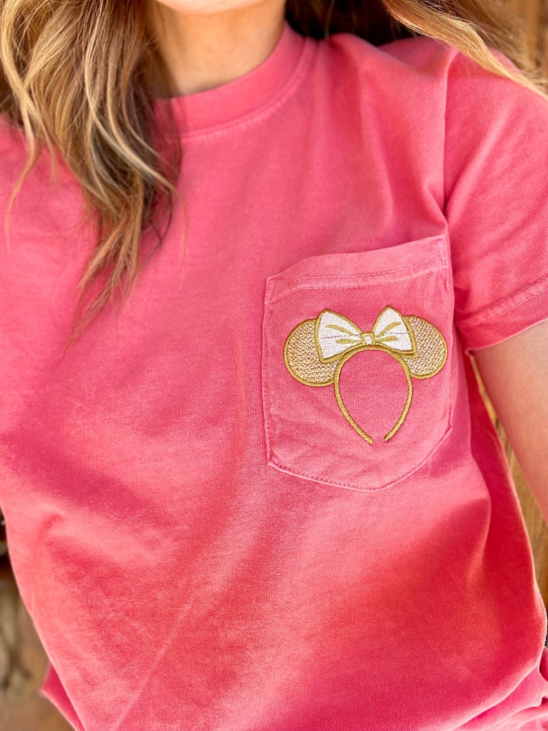 Op de afbeelding: Een roze T-shirt met een gouden geborduurde Mickey Mouse oren met een witte strik op een zak.