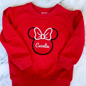 May include: Red sweatshirt with a white embroidered Minnie Mouse head and the name "Cecelia" underneath.
