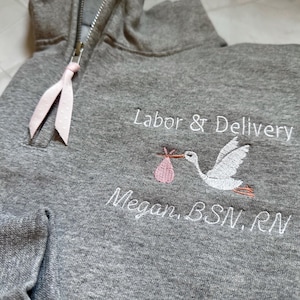 Puede incluir: Sudadera con capucha gris con cremallera a media altura y detalle de cinta rosa. La sudadera presenta texto bordado que dice "Labor & Delivery" y "Megan, BSN, RN" junto con una cig&uuml;e&ntilde;a que lleva un beb&eacute;.