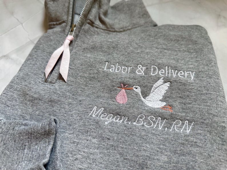 Puede incluir: Sudadera con capucha gris con cremallera a media altura y detalle de cinta rosa. La sudadera presenta texto bordado que dice "Labor & Delivery" y "Megan, BSN, RN" junto con una cig&uuml;e&ntilde;a que lleva un beb&eacute;.