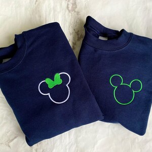 Op de afbeelding: Twee marineblauwe sweatshirts. E&eacute;n heeft een witte omtrek van Mickey Mouse met een groene strik, de andere heeft een groene omtrek van Mickey Mouse. Beide sweatshirts zijn opgevouwen en gepresenteerd op een wit oppervlak.