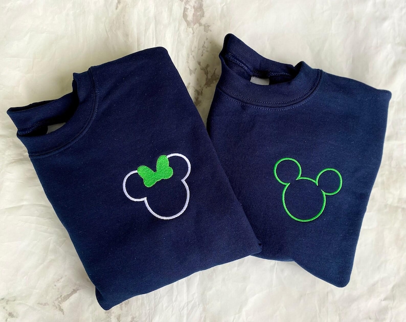 Op de afbeelding: Twee marineblauwe sweatshirts. E&eacute;n heeft een witte omtrek van Mickey Mouse met een groene strik, de andere heeft een groene omtrek van Mickey Mouse. Beide sweatshirts zijn opgevouwen en gepresenteerd op een wit oppervlak.
