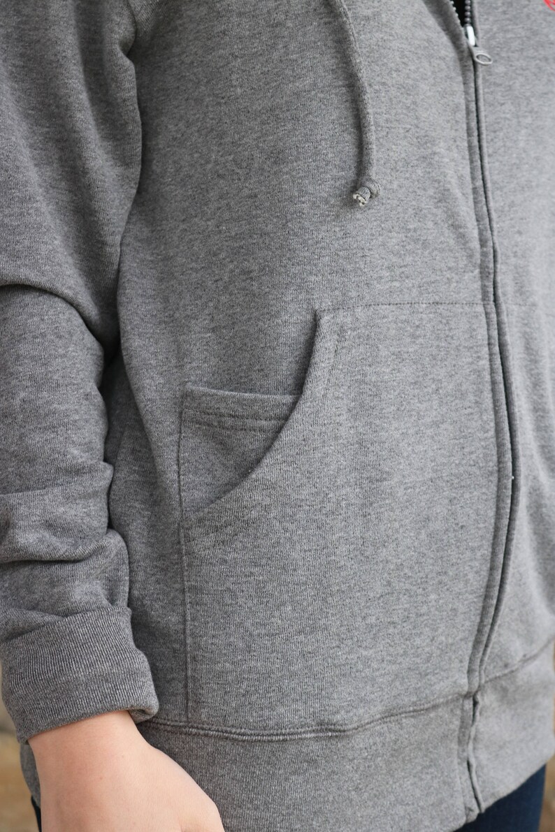 Puede incluir: Una sudadera con capucha gris con cremallera y un bolsillo canguro. La sudadera tiene una capucha con cord&oacute;n y mangas largas.