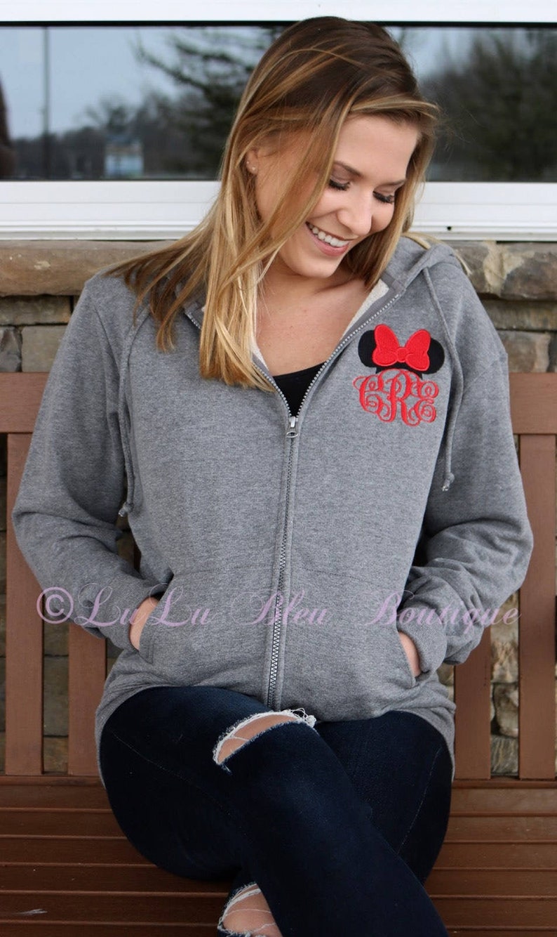 Puede incluir: Una sudadera gris con cremallera y capucha con un lazo rojo y un monograma "G" en la parte delantera. La sudadera la lleva puesta una persona con el pelo largo y rubio.