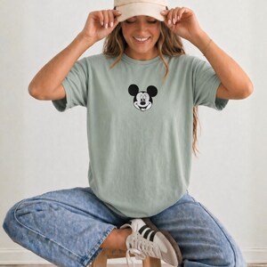 May include: A sage green short-sleeved t-shirt featuring a small black and white graphic of Mickey Mouse's face centered on the chest. The shirt has a crew neckline and a relaxed fit, paired with acid-wash blue denim jeans and white sneakers.