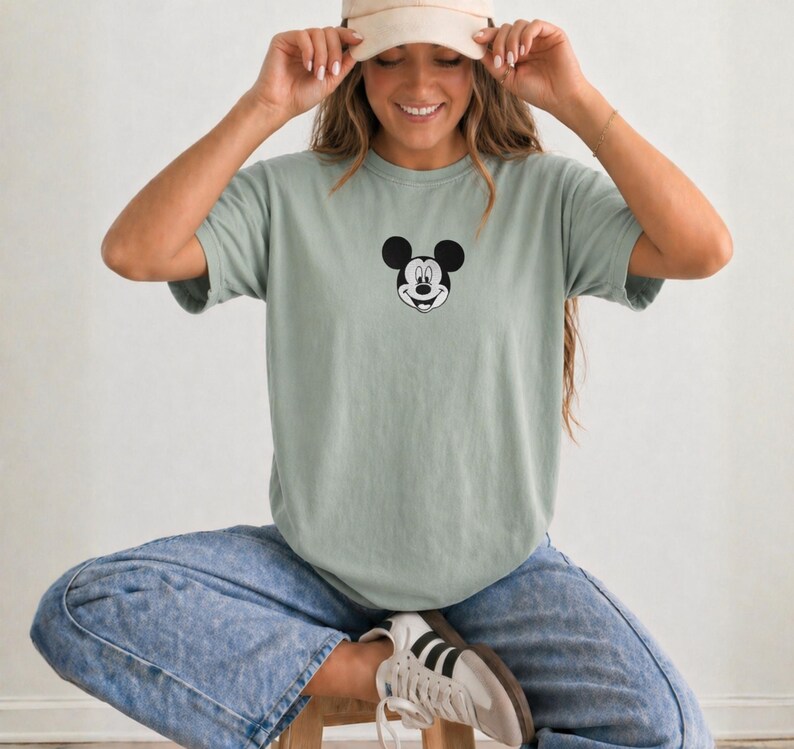May include: A sage green short-sleeved t-shirt featuring a small black and white graphic of Mickey Mouse's face centered on the chest. The shirt has a crew neckline and a relaxed fit, paired with acid-wash blue denim jeans and white sneakers.