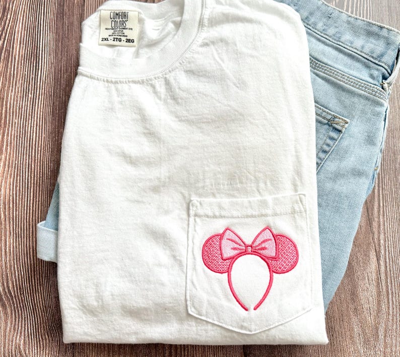 Op de afbeelding: Wit T-shirt met een roze geborduurd Minnie Mouse oren ontwerp op de zak. Het T-shirt heeft een label met de tekst "Comfort Colors" en "2XL - 2TG - 26G".