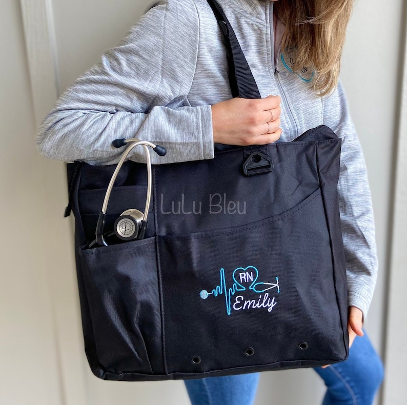 Puede incluir: Bolso tote negro con un estetoscopio en el bolsillo lateral. El bolso tiene un dise&ntilde;o bordado en azul de un coraz&oacute;n con "RN" dentro, una l&iacute;nea de latido del coraz&oacute;n y un estetoscopio. El texto "Emily" est&aacute; bordado debajo del dise&ntilde;o. El bolso tiene el texto "Lulu Bleu" impreso en la parte delantera.