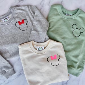 Op de afbeelding: Drie crewneck sweatshirts in grijs, gebroken wit en saliegroen. Elke sweatshirt heeft een zwarte Mickey Mouse-omtrek. De grijze sweatshirt heeft een rode strik, de gebroken witte heeft een roze strik en de groene heeft geen strik. Casual kleding.