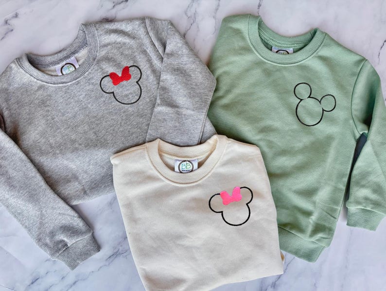 Op de afbeelding: Drie crewneck sweatshirts in grijs, gebroken wit en saliegroen. Elke sweatshirt heeft een zwarte Mickey Mouse-omtrek. De grijze sweatshirt heeft een rode strik, de gebroken witte heeft een roze strik en de groene heeft geen strik. Casual kleding.