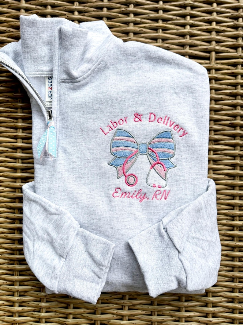 以下が含まれることがあります： 「Labor & Delivery」の刺繍が施された、リボンと聴診器のデザインが施されたライトグレーのクォータージップスウェットシャツ。名前「Emily, RN」が下に刺繍されています。ジッパーには、水色と白のドット柄のリボンが付いています。