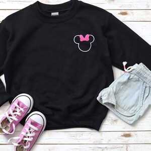Op de afbeelding: Een zwarte sweatshirt met een witte omtrek van een muizenkop en een roze strik. De afbeelding bevat ook lichtblauwe shorts en roze sneakers met witte veters, alles op een licht houten oppervlak.