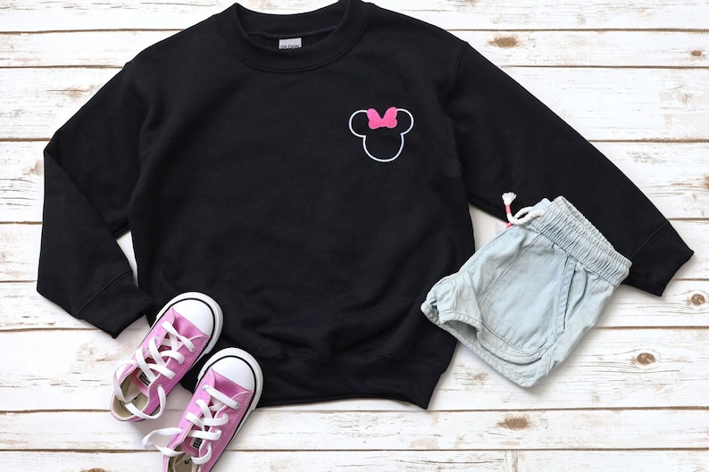Op de afbeelding: Een zwarte sweatshirt met een witte omtrek van een muizenkop en een roze strik. De afbeelding bevat ook lichtblauwe shorts en roze sneakers met witte veters, alles op een licht houten oppervlak.