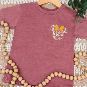 May include: A maroon short-sleeved t-shirt with a white daisy flower design in the shape of a mouse head with a yellow bow.