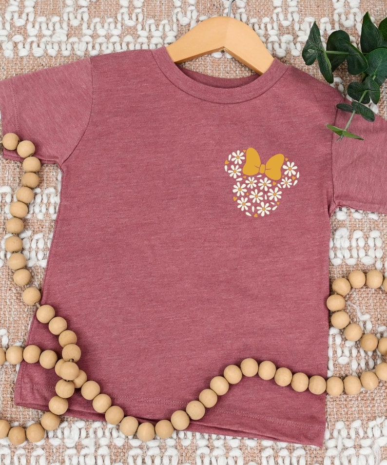 May include: A maroon short-sleeved t-shirt with a white daisy flower design in the shape of a mouse head with a yellow bow.
