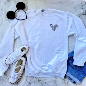 May include: White sweatshirt with a black and white leopard print Mickey Mouse head design on the front. The sweatshirt is laying on a white marble surface with a pair of white sneakers and a pair of black mouse ears.