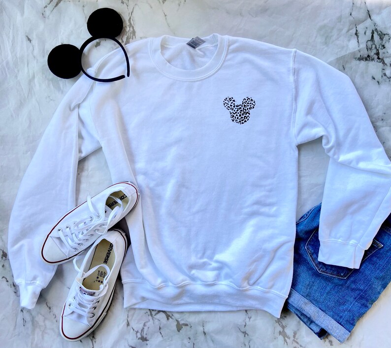 May include: White sweatshirt with a black and white leopard print Mickey Mouse head design on the front. The sweatshirt is laying on a white marble surface with a pair of white sneakers and a pair of black mouse ears.