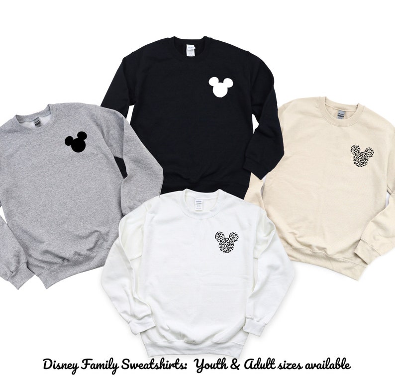 May include: Four sweatshirts with a Mickey Mouse silhouette on the front. The sweatshirts are in black, white, grey, and beige. The Mickey Mouse silhouette is black on the grey sweatshirt, white on the black sweatshirt, black with a white polka dot pattern on the white sweatshirt, and white with a black polka dot pattern on the beige sweatshirt. The text "Disney Family Sweatshirts: Youth & Adult sizes available" is at the bottom of the image.