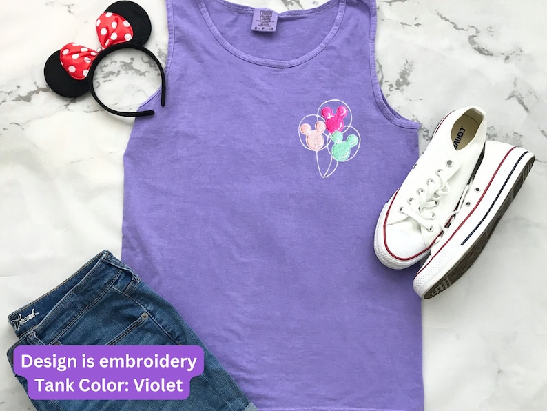 May include: A violet tank top with an embroidered design of three Mickey Mouse head balloons. The image also includes a pair of white sneakers, a Minnie Mouse ear headband, and a pair of blue denim shorts. The text on the image says "Design is embroidery" and "Tank Color: Violet".