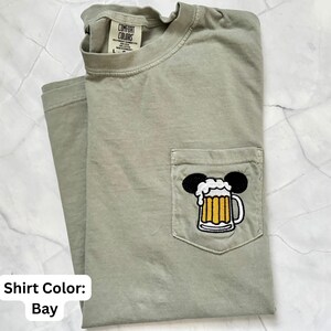 May include: A bay-colored short-sleeve t-shirt with a crew neckline and a chest pocket. The pocket features an embroidered graphic of a beer mug with white foam, topped with two black circular ears. The inner neck label reads Comfort Colors.