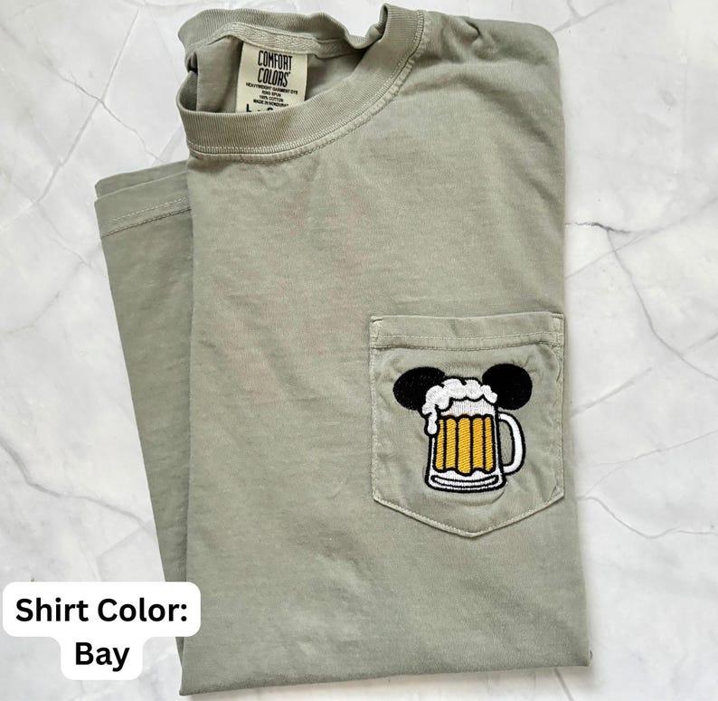 May include: A bay-colored short-sleeve t-shirt with a crew neckline and a chest pocket. The pocket features an embroidered graphic of a beer mug with white foam, topped with two black circular ears. The inner neck label reads Comfort Colors.