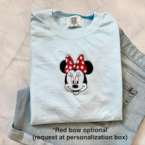 Peut inclure: Un t-shirt bleu clair avec un motif brod&eacute; de Minnie Mouse, orn&eacute; d'un n&oelig;ud rouge &agrave; pois blancs. Le t-shirt est pli&eacute; et plac&eacute; &agrave; c&ocirc;t&eacute; d'un short en jean bleu. Le t-shirt porte une &eacute;tiquette Comfort Colors.