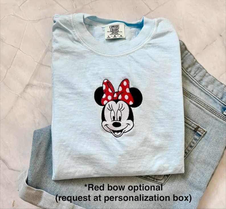 Peut inclure: Un t-shirt bleu clair avec un motif brod&eacute; de Minnie Mouse, orn&eacute; d'un n&oelig;ud rouge &agrave; pois blancs. Le t-shirt est pli&eacute; et plac&eacute; &agrave; c&ocirc;t&eacute; d'un short en jean bleu. Le t-shirt porte une &eacute;tiquette Comfort Colors.
