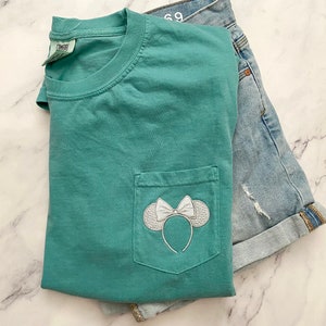 Op de afbeelding: Een tealblauw t-shirt met een wit geborduurd Minnie Mouse-oren en strik op de voorzak.