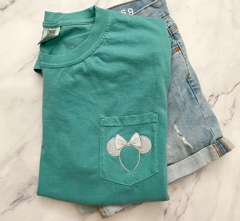 Op de afbeelding: Een tealblauw t-shirt met een wit geborduurd Minnie Mouse-oren en strik op de voorzak.