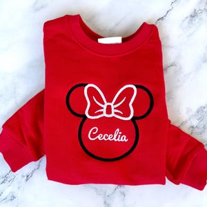 May include: Red sweatshirt with a black and white embroidered Minnie Mouse design and the name "Cecelia" underneath.