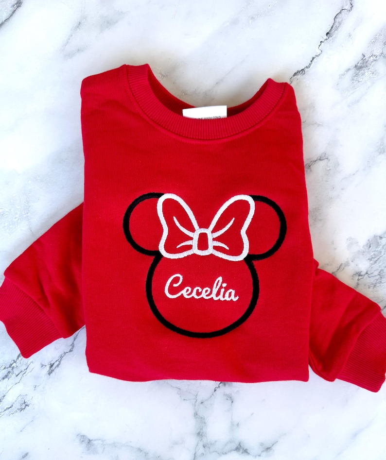 May include: Red sweatshirt with a black and white embroidered Minnie Mouse design and the name "Cecelia" underneath.