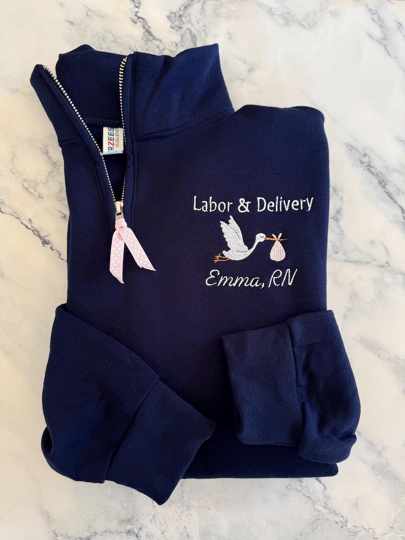 Puede incluir: Sudadera azul marino con cremallera de cuarto de cierre y cig&uuml;e&ntilde;a bordada que lleva un beb&eacute;. El texto "Labor & Delivery" y "Emma, RN" est&aacute;n bordados debajo. Una cinta rosa est&aacute; sujeta a la cremallera.