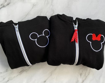May include: Two black hoodies with white embroidered Mickey Mouse and Minnie Mouse designs. The Minnie Mouse hoodie has a red polka dot ribbon on the zipper.