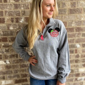 Op de afbeelding: Een grijs sweatshirt met een zwart en rood appelontwerp met een monogram. De appel heeft een groen blad en een zwart Mickey Mouse-oor aan elke kant.