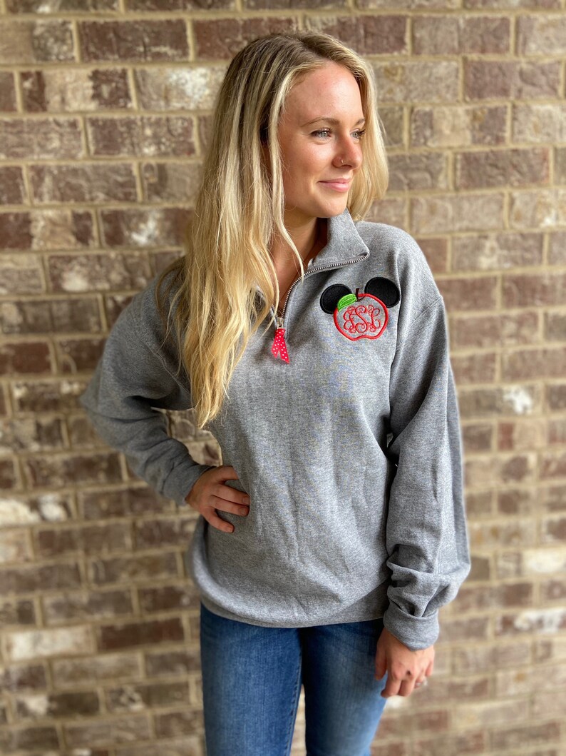 Op de afbeelding: Een grijs sweatshirt met een zwart en rood appelontwerp met een monogram. De appel heeft een groen blad en een zwart Mickey Mouse-oor aan elke kant.