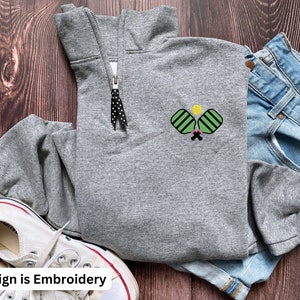 May include: Grey sweatshirt with a black zip and an embroidered design of two pickleball paddles and a ball in green and yellow. The design is on the left side of the sweatshirt.