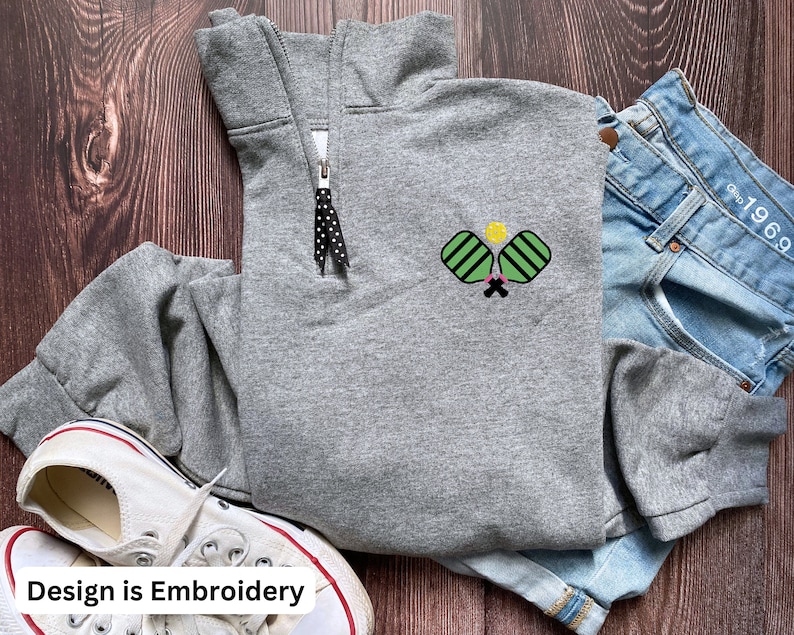 May include: Grey sweatshirt with a black zip and an embroidered design of two pickleball paddles and a ball in green and yellow. The design is on the left side of the sweatshirt.