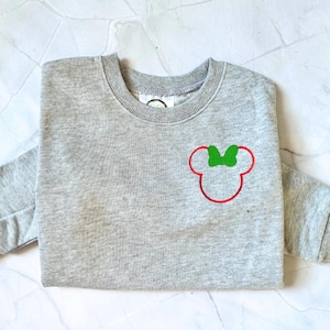 Op de afbeelding: Een grijze sweatshirt met een rood en groen Minnie Mouse-ontwerp. Het ontwerp staat op de voorkant. De sweatshirt heeft lange mouwen en een ronde hals.