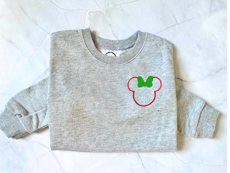 Op de afbeelding: Een grijze sweatshirt met een rood en groen Minnie Mouse-ontwerp. Het ontwerp staat op de voorkant. De sweatshirt heeft lange mouwen en een ronde hals.