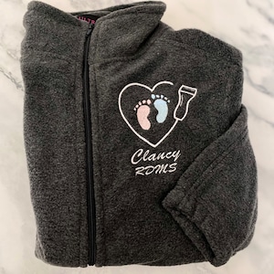 Könnte beinhalten: Eine graue Fleecejacke mit einem weißen gestickten Herzdesign mit zwei Baby-Fußabdrücken, einem rosa und einem blauen. Der Text "Clancy RDMS" ist unter dem Herzen gestickt.