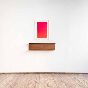 May include: A framed red and orange gradient print hangs above a wooden shelf against a white wall. The floor is made of light wood planks.