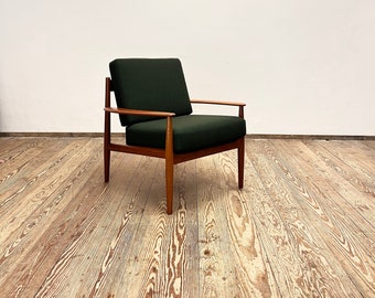 May include: A dark green armchair with wooden legs and arms sits on a light wood floor. A red framed picture hangs on the white wall behind the chair.