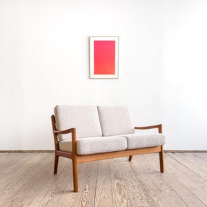 Puede incluir: Un sof&aacute; de dos plazas de estilo moderno de mediados de siglo, con una tela clara y texturizada y un marco de madera. El sof&aacute; tiene un dise&ntilde;o minimalista. Una obra de arte enmarcada con un degradado de rosa y rojo cuelga de la pared blanca detr&aacute;s del sof&aacute;.