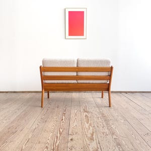 Puede incluir: Un sof&aacute; de dos plazas de estilo moderno de mediados de siglo, con estructura de madera clara y cojines beige. El sof&aacute; est&aacute; colocado contra una pared blanca, con una obra de arte enmarcada que muestra un degradado de rosa y rojo. La habitaci&oacute;n tiene suelo de madera.