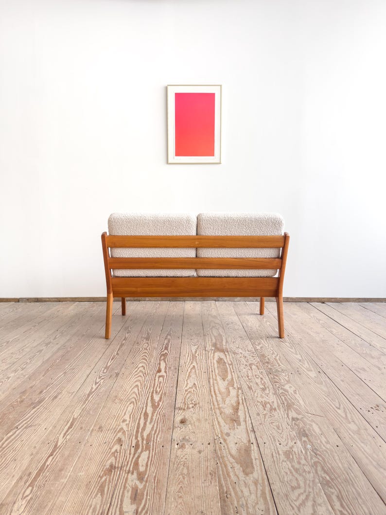 Puede incluir: Un sof&aacute; de dos plazas de estilo moderno de mediados de siglo, con estructura de madera clara y cojines beige. El sof&aacute; est&aacute; colocado contra una pared blanca, con una obra de arte enmarcada que muestra un degradado de rosa y rojo. La habitaci&oacute;n tiene suelo de madera.